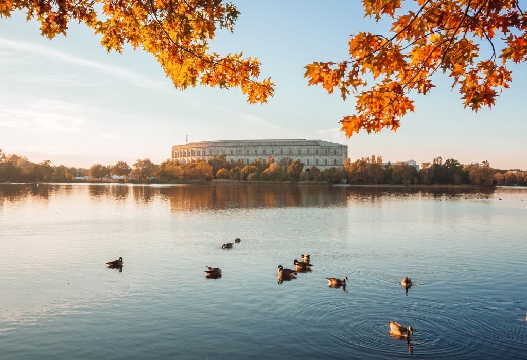 A lake with ducks and fall foilage in the foreground and the round former NAZI rally grounds (now a museum) in the background