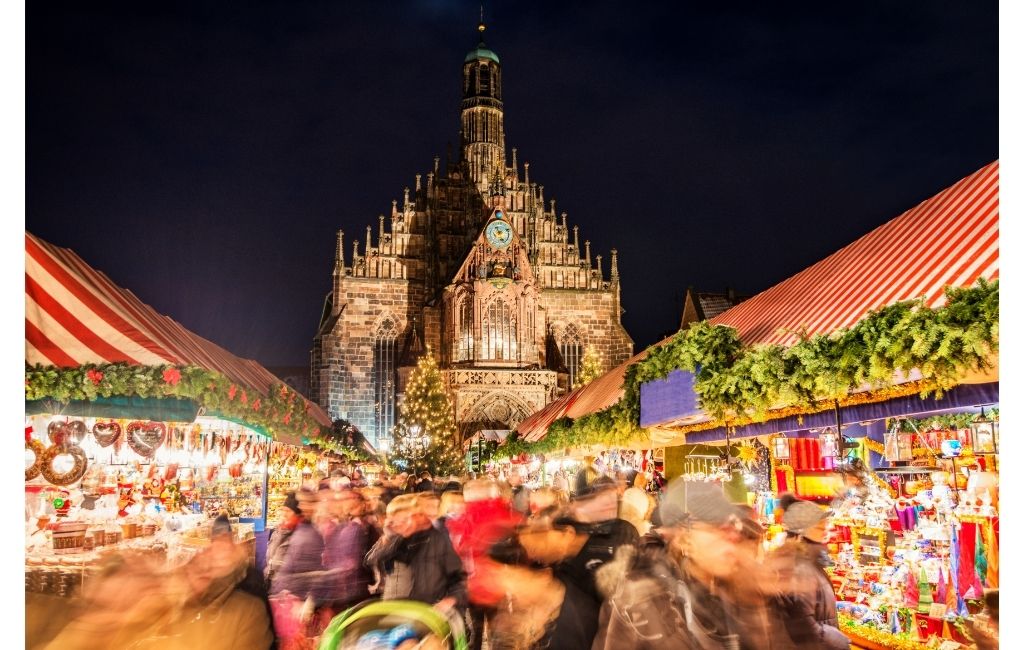 Nuremberg Christmas Market with Christmas market booths on both sides, a church in the background and people in a stream in the middle of the photo